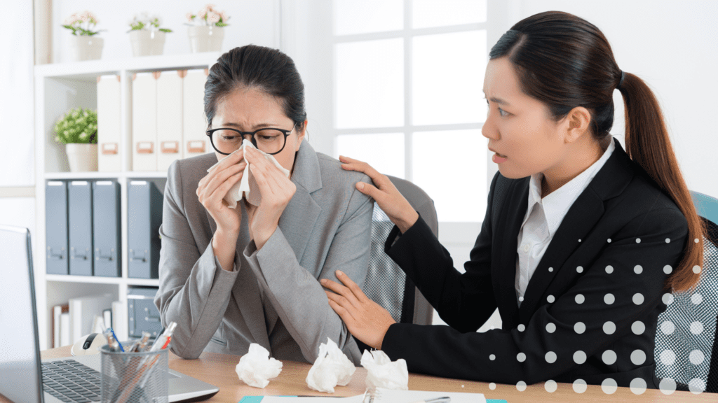 A woman in glasses blows her nose while another woman comforts her. Both are in suits in an office setting.